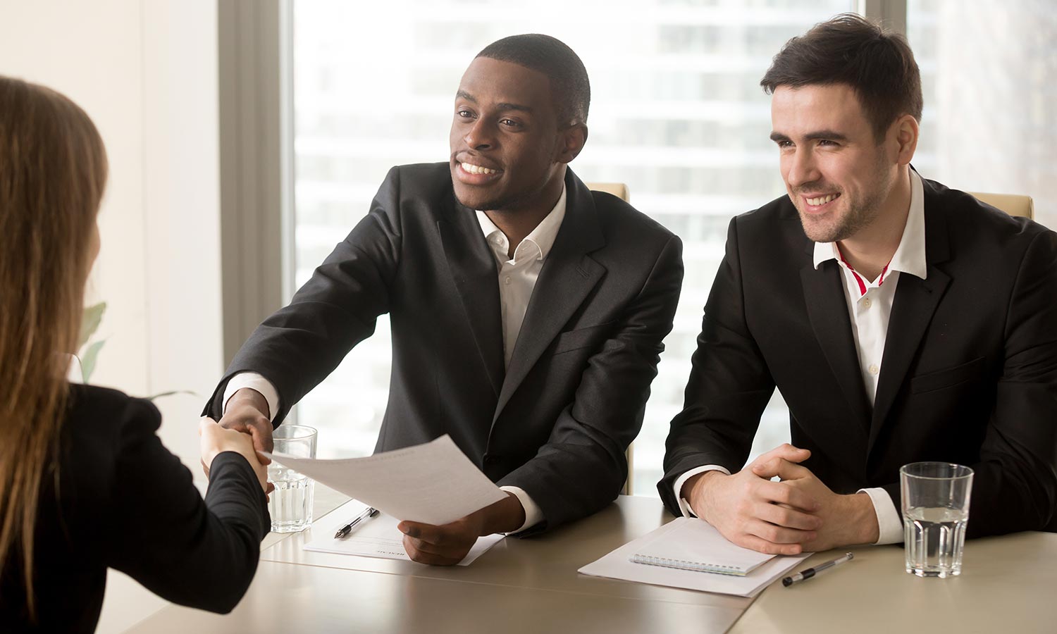 Two cheerful black and white recruiters welcoming female applicant on job interview, african and caucasian hr managers greeting candidate for vacant position, handshaking and good first impression